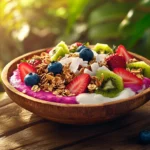 A vibrant pitaya bowl topped with fresh fruits, granola, and coconut flakes on a rustic wooden table with tropical plants in the background